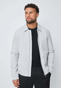 Homme aux cheveux bouclés courts et à la barbe, portant une veste à zip gris clair sur une chemise noire et un pantalon noir, les mains dans les poches, regardant vers la droite.