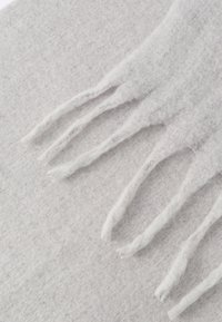 Svetlosivý šál s jemným, textúrovaným povrchom. Má dlhé, rozstrapkané okraje a nenápadné variácie farby a hĺbky po celom materiáli.