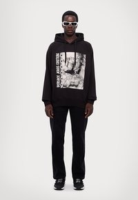 Felpa nera oversize con una stampa grafica, testo bianco di lato, abbinata a pantaloni neri e sneakers nere. Il modello indossa occhiali da sole bianchi.