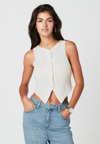 Biały, bezrękawnikowy crop top z dzianiny o prążkowanej fakturze, z dekoltem w kształcie litery V i guzikami z przodu. W komplecie z jasnoniebieskimi spodniami typu high-waisted.