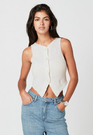 Bílý, bezrukávový pletený crop top s žebrovanou texturou, se V-výstřihem a předními knoflíky. V kombinaci s džínami světle modré barvy s vysokým pasem.