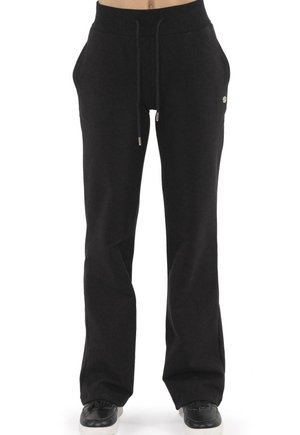 Modèle portant un pantalon de jogging noir taille haute et à jambes larges, avec un cordon de serrage, et des baskets noires sur fond blanc.