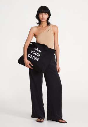 Borsa tote nera con la scritta "IO SONO TUA SORELLA" in bianco, abbinata a un top beige a spalla unica e pantaloni neri larghi. Sandali piatti visibili.