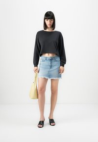 Young woman with black long-sleeve cropped top, light blue denim mini skirt, black open-toe heels, and a beige handbag standing against a white wall.