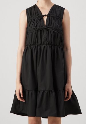 Robe noire sans manches avec des volants froncés et deux nœuds noués sur la poitrine, portée par une personne debout sur un fond uni.