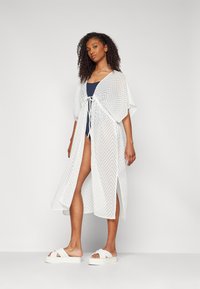 Cover-up bianco e leggero con un motivo a zigzag, apertura frontale, maniche corte e chiusura con lacci, indossato sopra un costume da bagno blu navy con grandi spacchi.