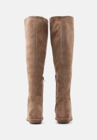 Ζευγάρι ψηλές σουέντ μπότες χρώματος ταουπέ με χαμηλά ξύλινα τακούνια, εμφανιζόμενες από πίσω σε λευκό φόντο.