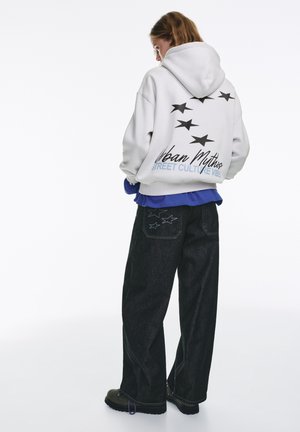 Fehér kapucnis pulóver fekete csillag grafikával és felirattal a hátán, kék felső rétegében; sötét farmernadrággal párosítva, mely csillag hímzéssel díszített.