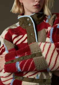 Veste en polaire rouge, crème et marron avec un motif géométrique ; possède un col montant et des passepoils bleus ; fermetures à boutons sur le devant.