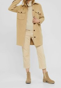 Kvinna klädd i en beige knappad lång kappa, ljus polotröja, cropped byxor och tanfärgade ankelstövlar mot en enkel bakgrund.