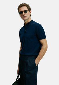 Polo shirt bleu marine à manches courtes avec un col et deux boutons, associé à un pantalon sombre, tenant un sac dans une main, en tissu texturé.