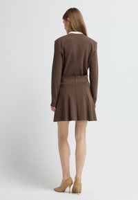Bruine jurk met een fitted lijfje en uitlopende rok, voorzien van een zichtbare rits op de rug. Geaccentueerd met een witte kraag en gecombineerd met beige enkellaarzen.