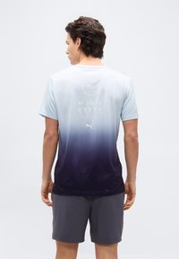 Άνδρας με την πλάτη, που φοράει αθλητικό μπλουζάκι με βαθμιδωτό μπλε χρώμα και το κείμενο «My Sport is HYROX» και σκούρο σορτς σε μονόχρωμο λευκό φόντο.