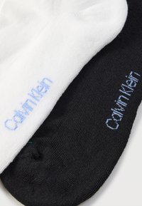 Δύο βαμβακερές κάλτσες αστραγάλου, μία λευκή και μία μαύρη, καθεμία με το κέντημα "Cottonrich" σε ανοιχτό μπλε κοντά στην περιοχή της μανσέτας.