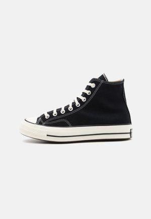 Black canvas high-top sneaker with white laces, rubber toe cap, and a contrasting white sole featuring black stripes along the edge.