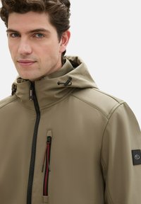 Lightweight beige jacket featuring a zippered chest pocket with red accent, drawstring hood, and textured fabric. Black logo patch on sleeve.