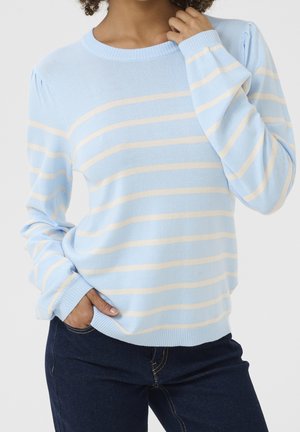 Vrouw draagt een lichtblauwe trui met crèmekleurige horizontale strepen en donkerblauwe jeans, poseert met één hand in haar zak en de andere dicht bij haar hals.