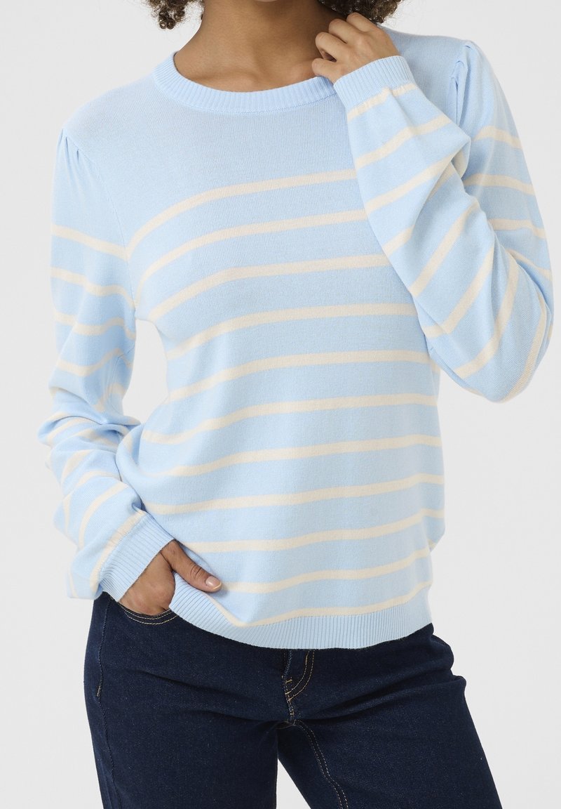 Femme portant un pull bleu clair avec des rayures horizontales crème et un jean bleu foncé, posant avec une main dans la poche et l'autre près du cou.