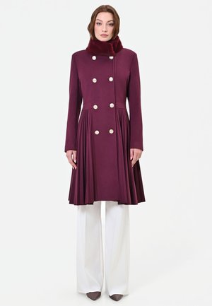 Donna che indossa un lungo cappotto doppiopetto bordeaux con gonna plissettata e collo in pelliccia, abbinato a pantaloni bianchi a gamba larga e scarpe marroni.