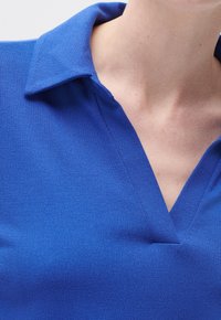 Blå stickad polo med en texturerad yta, med V-ringning och en mjuk, strukturerad krage. Tyget verkar lätt och andningsbart.