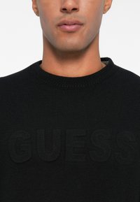 Maglione nero a maglia con colletto girocollo, con un logo "GUESS" testurizzato incorporato in modo sottile nel tessuto. Texture liscia, design casual.