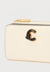 Cartera de cuero beige con cremallera dorada y emblema "C" en negro y dorado en la parte frontal, sobre fondo blanco.