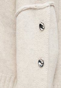 Tissu tricoté couleur crème avec deux boutons métalliques ovales brillants disposés verticalement près du rabat d'une poche avant.