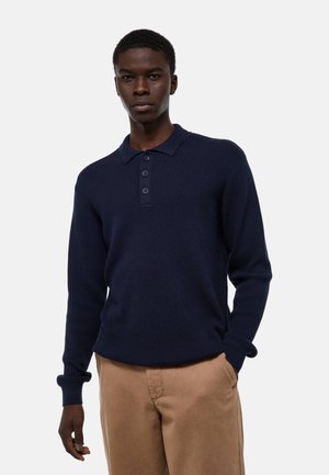 Jeune homme portant un polo à manches longues en maille bleu marine et un pantalon beige, debout avec une main dans la poche contre un fond uni.