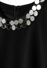 Zwarte stoffen top met een ritssluiting, voorzien van een ronde kraag versierd met reflecterende, ronde pailletten in een patroon.