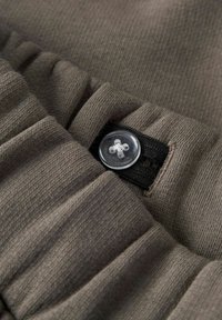 Ceinture élastique réglable en tissu tricoté marron, maintenue par un bouton transparent sur une bande élastique noire.