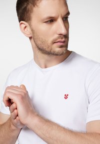 Biała koszulka z okrągłym dekoltem, wykonana z bawełny, z małym czerwonym haftowanym logo na piersi. Gładka tekstura, dopasowany krój.