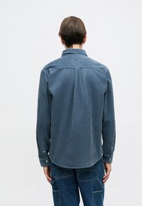 Pánska kordurová košeľa v tlmenom modrom odtieni, s vertikálnou ribovanou textúrou, špicatým golierom a gombíkovými manžetami, s zaobleným lemom na zadnej strane.