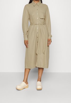 Woman wearing a beige, long-sleeve, button-down midi dress with a front pocket and matching belt, paired with cream platform clogs.