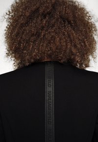 Černý oděv s vysokým výstřihem, vyrobený z texturované tkaniny. Po zadní straně vede vertikální černý pás s brandingem "Karl Lagerfeld".