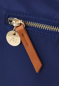 Námornícky látkový sáček s zlatým zipem, hnědým koženým táhlíkem na zip a kulatým zlatým přívěskem s abstraktním designem.