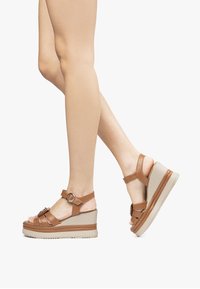 Sandali con zeppa marroni caratterizzati da una fascia testurizzata, dettaglio con fibbia e una suola piatta beige. Lo stile open-toe mette in mostra i piedi.