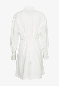 Vit långärmad klänning med samlad elastisk midja och knäppta ärmslut, visad från ryggen mot en neutral bakgrund.