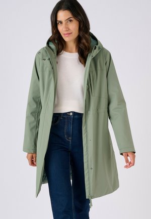 Abrigo impermeable largo verde con capucha, con cierres de presión, dos bolsillos delanteros y corte recto. Combinado con vaqueros azules.