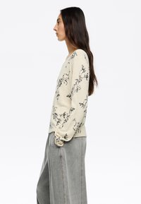 Pull léger beige avec un motif floral noir, décolleté rond et manches longues, associé à un pantalon large gris. Texture riche et matière douce.