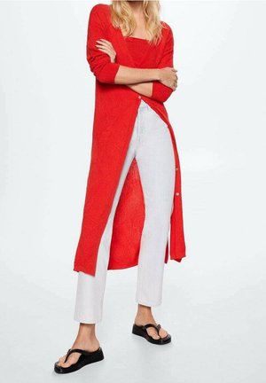 Femme portant un long cardigan rouge, un jean blanc et des sandales plateformes noires, debout les bras croisés sur un fond blanc.
