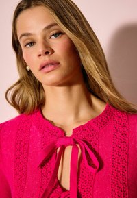 Bluză tricotată roz strălucitor, cu decolteu rotund, detalii intricate asemănătoare dantelei și accent de fundiță în față. Textură netedă.