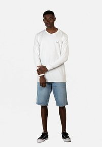 Weißes Langarmhemd mit kleinem schwarzem Logo auf der Brust, kombiniert mit hellblauen Jeansshorts und schwarzen Sneakers.