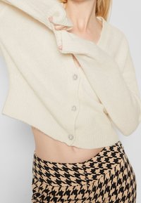 Krémový zkrácený kardigan s ribbovanou texturou, s pěti stříbrnými knoflíky. Spárovaný s sukní v houndstooth vzoru.