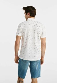 Biała koszulka polo z wzorem małych czarnych strzałek, krótki rękaw i kołnierzyk. W połączeniu z niebieskimi szortami dżinsowymi.