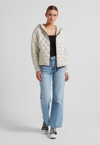 Gepolsterte Cremejacke mit Kapuze, diamantförmigem Steppmuster, schwarzem Reißverschluss und gerippten Bündchen, kombiniert mit hellblauen Schlagjeans und schwarzen Sneakers.