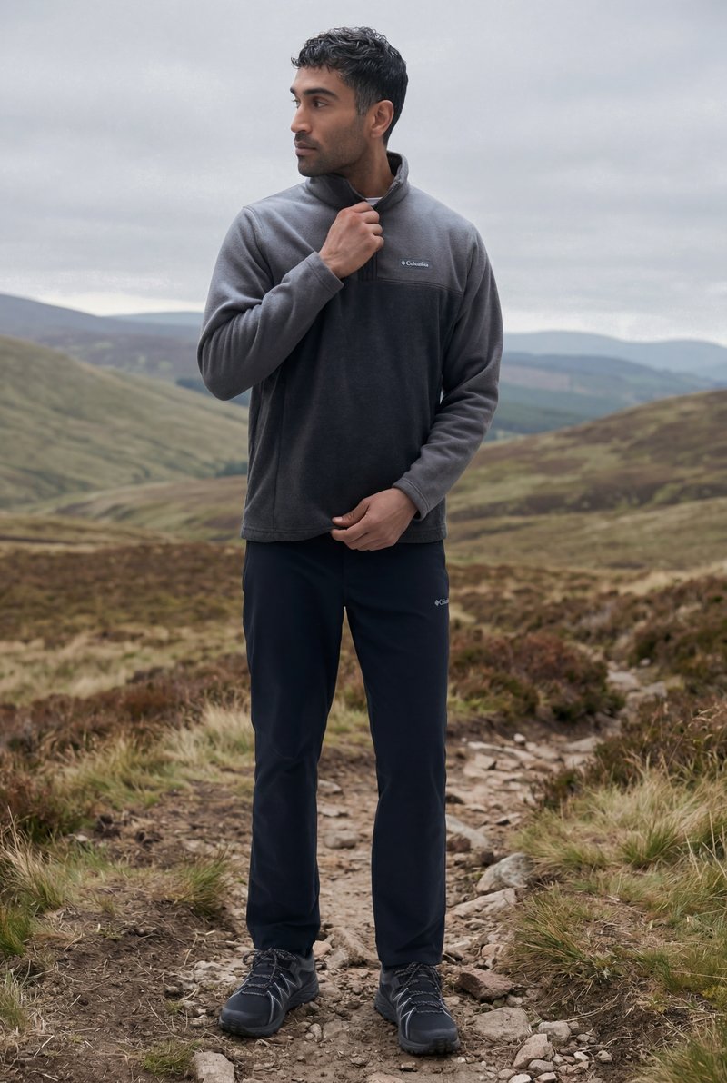 Bărbat ajustând o geacă fleece Columbia gri, stând pe un traseu stâncos într-un peisaj învăluit în dealuri, sub un cer înnorat.