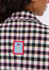 Žena nosí černou, bílou, růžovou a žlutou kostkovanou bundu s jasným nášivkem na zadní straně, které říká "To nejlepší teprve přijde".