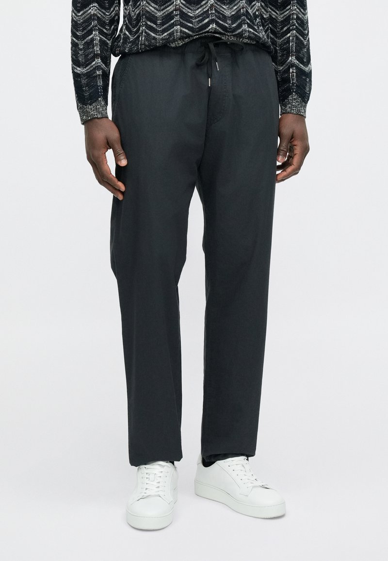 Pantalon fuselé gris foncé, léger, avec une taille élastique et un cordon de serrage, associé à des baskets blanches. Texture lisse, design minimaliste.