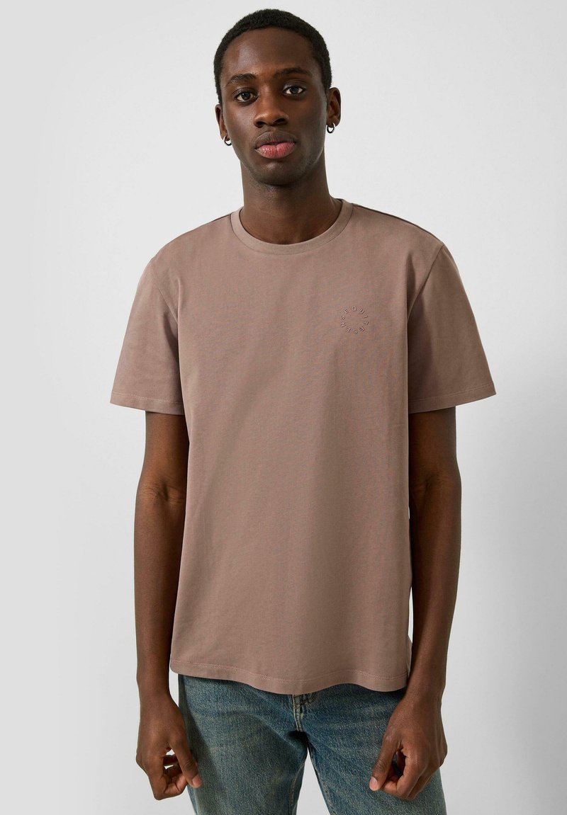 T-shirt à manches courtes, col rond, en coton marron terreux, avec un logo circulaire discret sur la poitrine gauche. Porté avec un jean denim bleu.