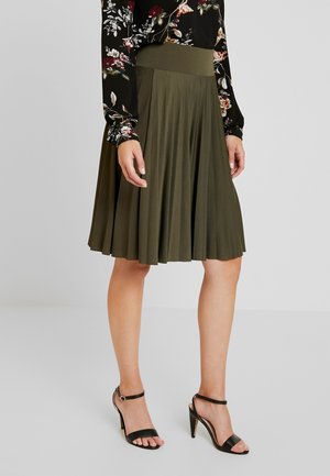 Kvinde iført plisseret olivengrøn knælængde nederdel, blomsterprikket bluse med lange ærmer og sorte høje hæle, stående på gråt gulv.